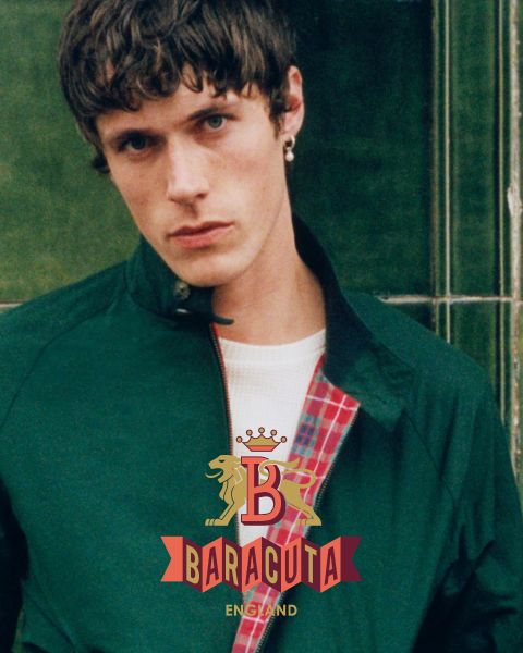 Una persona con cabello corto y oscuro y un pendiente, que lleva una chaqueta oscura sobre una camisa blanca, mira a la cámara. El interior de la chaqueta es de cuadros rojos. En la parte delantera se muestra el logotipo de Baracuta con un león y una "B".