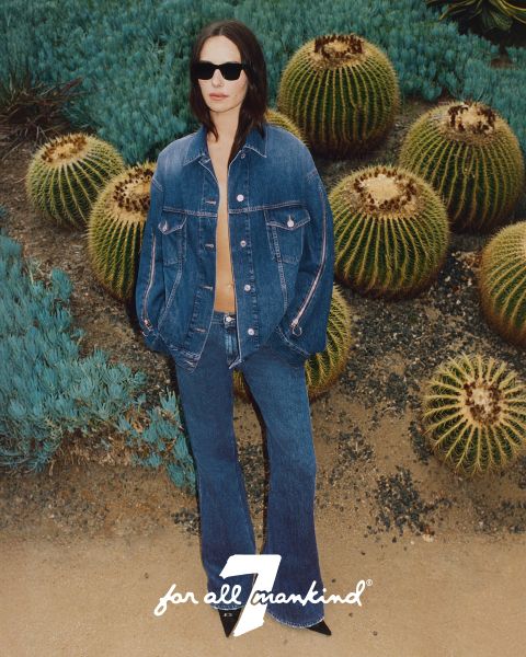 Una persona con chaqueta y pantalones vaqueros está de pie entre cactus. Lleva gafas de sol y la chaqueta está ligeramente abierta. El suelo es arenoso y al fondo se ven suculentas verdes. Abajo se ve el logo "7 for all mankind".