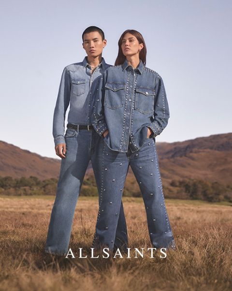 Dos personas están de pie en un campo abierto, vestidas con conjuntos de mezclilla adornados con remaches. La persona de la izquierda lleva una camisa de mezclilla clara y vaqueros, mientras que la persona de la derecha lleva una chaqueta de mezclilla oscura y vaqueros. Abajo pone "ALLSAINTS".
