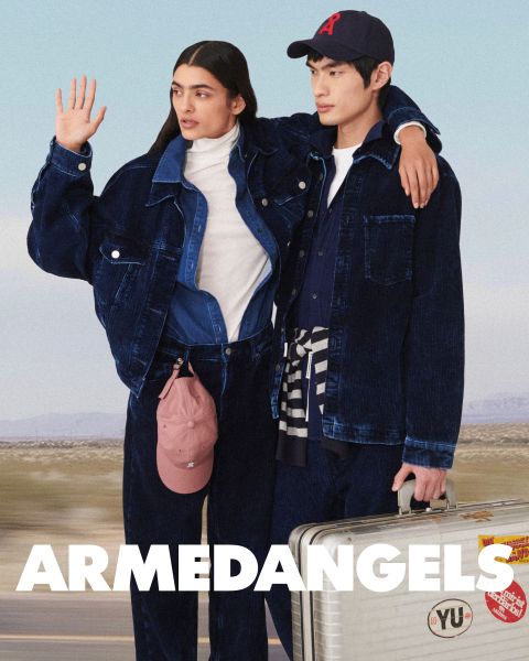 Dos jóvenes adultos vestidos con ropa vaquera están juntos al aire libre, uno con el brazo levantado, el otro con una maleta cubierta de pegatinas. En la parte inferior, en letras blancas y negrita, dice "ARMEDANGELS".