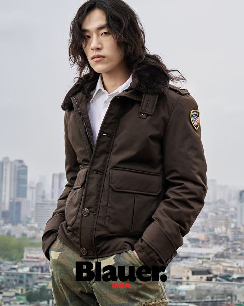 Una persona con pelo largo, que lleva una chaqueta marrón y pantalones de camuflaje, está de pie frente a un paisaje urbano. La chaqueta tiene un parche en el hombro y abajo se puede ver la palabra "Blauer". El cielo está nublado.