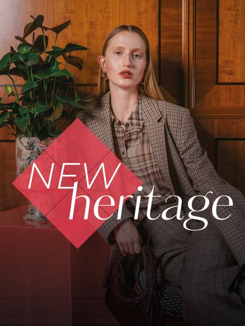 Una mujer con un traje a cuadros está sentada junto a una planta, con las palabras "NEW heritage" superpuestas frente a ella.