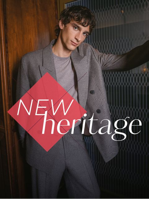 Un hombre con un traje gris está de pie frente a una pared de madera con las palabras "NEW heritage" en negrita.