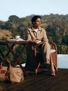 Una persona con ropa ligera está sentada descalza en una mesa con café, al aire libre junto a la piscina, con árboles y colinas de fondo. Al lado de la mesa hay un bolso.