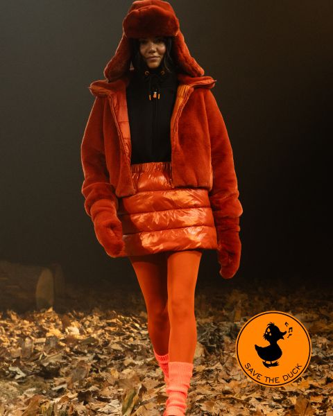 Una modelo camina por un sendero cubierto de hojas. Lleva una chaqueta de piel naranja, una falda a juego y un gorro con medias naranjas y calcetines color melocotón. En la esquina se ve el logo de "Save the Duck".