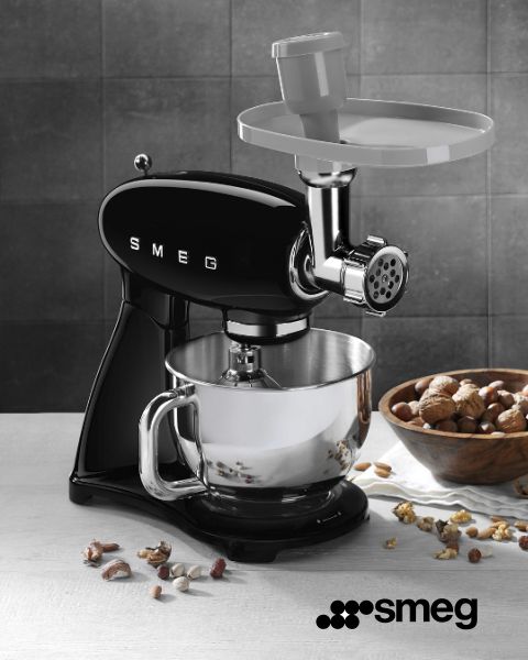 Una batidora Smeg negra con un accesorio para picar carne está sobre una encimera de cocina. Debajo hay un cuenco mezclador plateado. Nueces enteras y picadas están esparcidas sobre una superficie de madera cercana. En la esquina inferior derecha se ve el logotipo de Smeg.