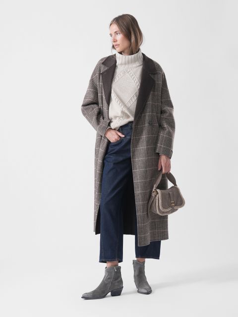 Una mujer con un abrigo a cuadros, un suéter, jeans oscuros y botas grises sostiene un pequeño bolso; está de pie mirando hacia la izquierda frente a un fondo blanco liso.