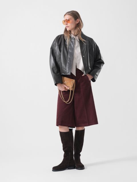 Una mujer con una chaqueta de cuero negra, un culotte burdeos, botas hasta la rodilla y gafas de sol, sosteniendo un bolso marrón y de pie frente a un fondo blanco liso.