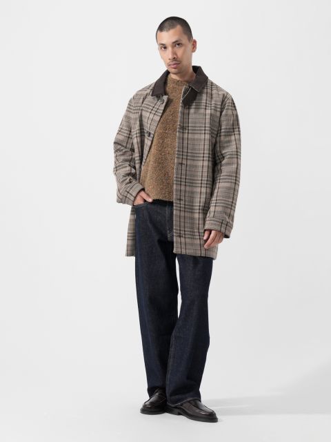Un hombre con un abrigo a cuadros, un suéter marrón, jeans oscuros y zapatos negros, de pie frente a un fondo blanco liso.