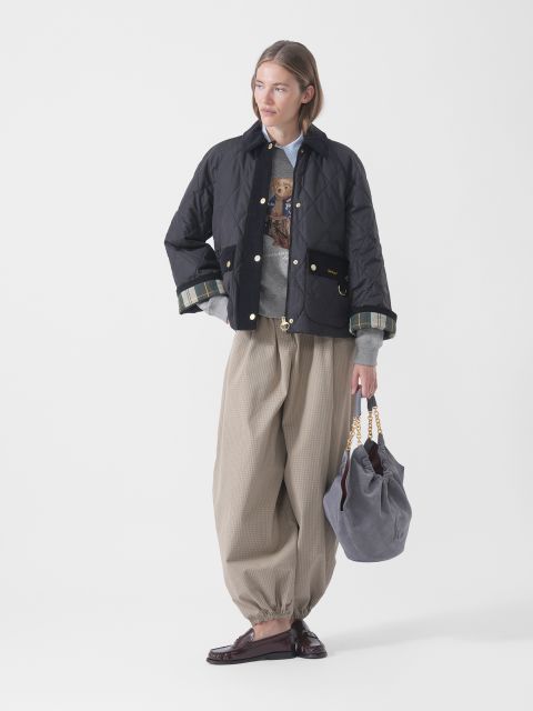 Una persona con una chaqueta acolchada negra, un suéter gris, pantalones anchos beige y mocasines sostiene un bolso gris con asas de cadena doradas y está de pie frente a un fondo blanco.