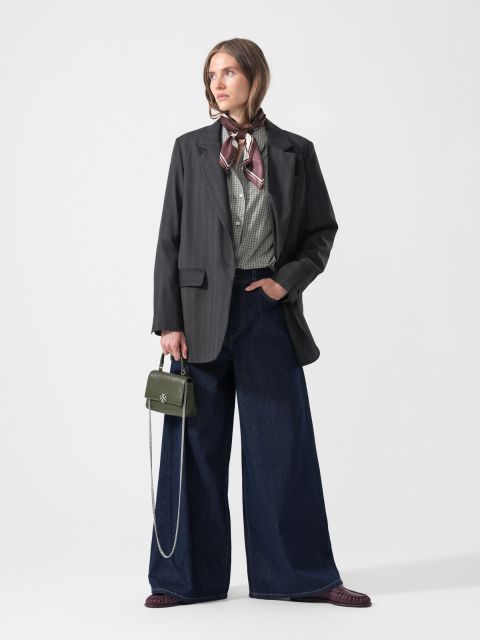 Una mujer en jeans de pierna ancha, una camisa a cuadros, un blazer oscuro y una bufanda, sosteniendo un pequeño bolso verde, de pie frente a un fondo blanco liso.