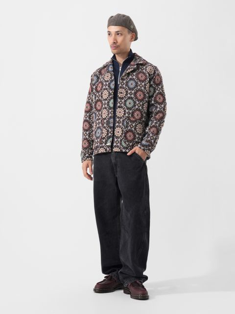 Un hombre con una chaqueta estampada, pantalones negros, zapatos granates y una boina gris posa frente a un fondo blanco liso.
