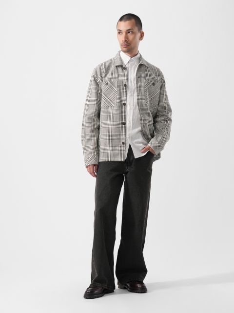 Sobre un fondo blanco liso, un hombre lleva una chaqueta a cuadros sobre una camisa blanca, pantalones negros holgados y zapatos negros.
