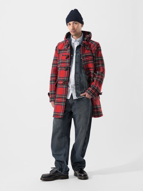 Una persona con un abrigo de cuadros rojo, una chaqueta vaquera, una camisa blanca, vaqueros anchos, zapatos negros y un gorro de punto oscuro, de pie frente a un fondo blanco liso.