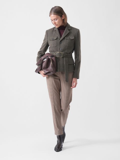 Una mujer con un blazer con cinturón, pantalones a cuadros y botas sostiene un bolso marrón, mira hacia abajo y se para frente a un fondo blanco liso.