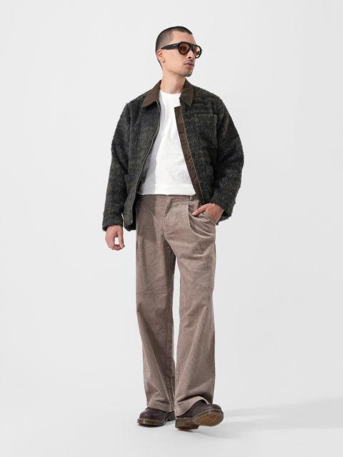 Un hombre con gafas de sol, una chaqueta oscura y peluda, una camiseta blanca, pantalones anchos de color tostado y zapatos marrones posa frente a un fondo blanco liso.