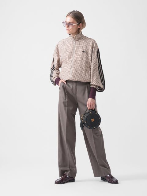 Una persona con gafas de sol lleva una chaqueta a cuadros, pantalones anchos grises y zapatos burdeos, y sostiene un pequeño bolso redondo negro delante de un fondo blanco liso.
