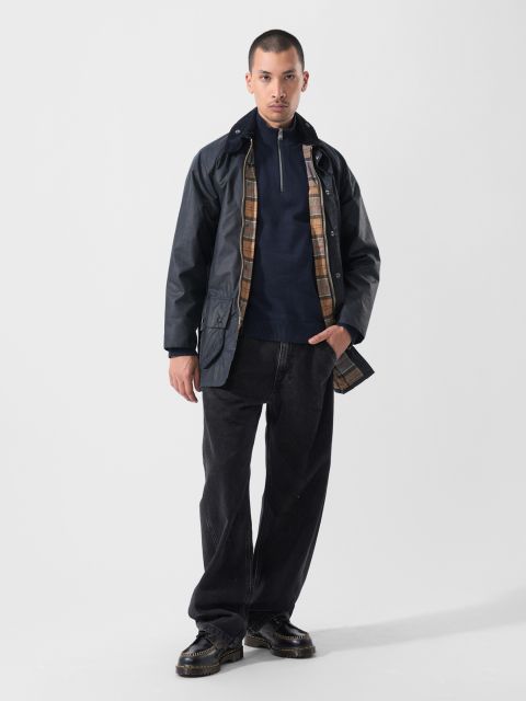 Un hombre con ropa informal de pie frente a un fondo blanco liso: chaqueta oscura con forro a cuadros, jersey azul marino con cremallera, vaqueros negros holgados y zapatos negros.
