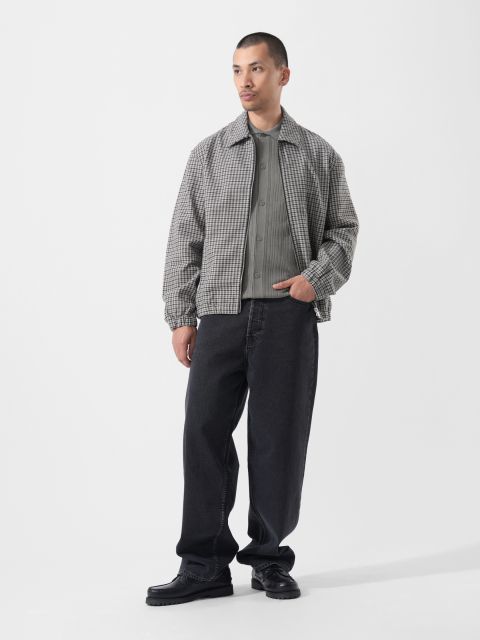 Sobre un fondo blanco liso, un hombre lleva una chaqueta de cuadros gris, una camisa gris, vaqueros negros holgados y zapatos negros.