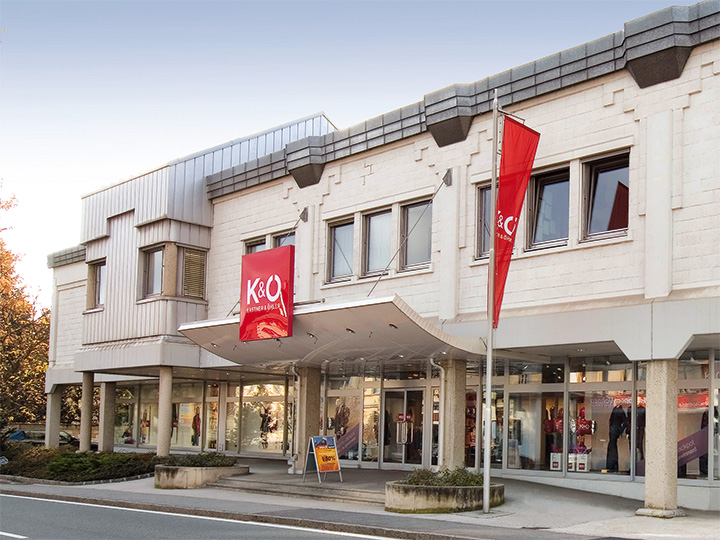 El moderno edificio con fachada beige alberga las tiendas K&Ö Kastner & Öhler y Giga X Sport. Los letreros rojos y naranjas son llamativos. Grandes ventanas y una calle delante bajo un cielo despejado completan la imagen.