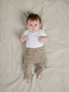 Un bebé sonriente está acostado sobre una manta blanca, vistiendo un suéter de punto azul, pantalones de pana marrones con bolsillos y calcetines grises. El bebé parece feliz y relajado, con los brazos ligeramente doblados a la altura de los codos.