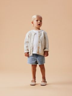 Una joven sonriente con un chándal color lavanda de pie frente a un fondo gris, con un lazo blanco muy grande en la cabeza.