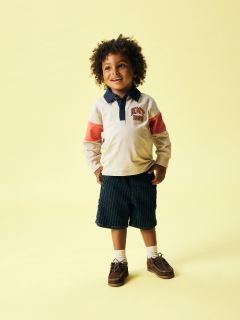Un chico con cabello rubio lleva una gorra negra, una chaqueta de forro polar color crema sobre una camisa a rayas, vaqueros grises y zapatillas de deporte. Está de pie frente a un fondo azul liso y mira hacia abajo con las manos juntas.