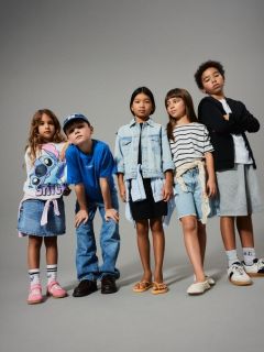 Una niña pequeña con cabello rubio está de pie sonriendo frente a un fondo verde, vistiendo un suéter marrón sin mangas sobre un vestido floreado, calcetines hasta la rodilla y zapatos marrones.