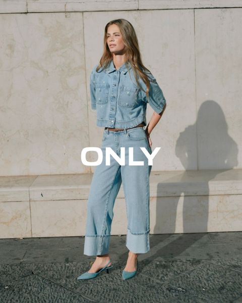 Una joven con un abrigo negro, una camisa a rayas azules y una falda marrón rojizo camina por una calle de la ciudad con edificios históricos al fondo. La palabra "ONLY" se muestra en la imagen.