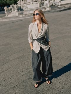 Una mujer con un traje marrón de gran tamaño, cinturón negro y joyería de oro camina con confianza por una calle de la ciudad, llevando un bolso negro grande. En el fondo se ven personas sentadas en un café.