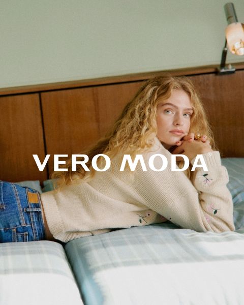 Una mujer con gafas y un vestido floreado con volantes se apoya en una pared blanca frente a un fondo de azulejos azules. La inscripción "VERO MODA" aparece en el centro de la imagen.