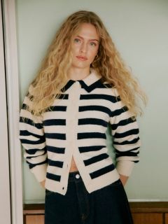 Una mujer con cabello rizado está de pie junto a una pared de azulejos. Lleva un cárdigan de punto gris claro y esponjoso, pantalones cortos de cuero negros de talle alto y medias negras, con las manos en los bolsillos y una expresión facial segura.