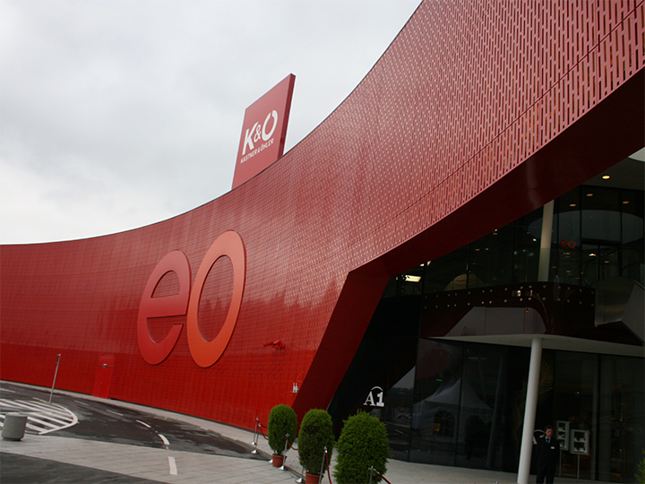Un edificio moderno con una gran fachada roja curvada con las letras "eo". En el techo se ven letreros de K&O y Giga Sport. La entrada es de cristal, con plantas en macetas al lado, y una curva en primer plano.