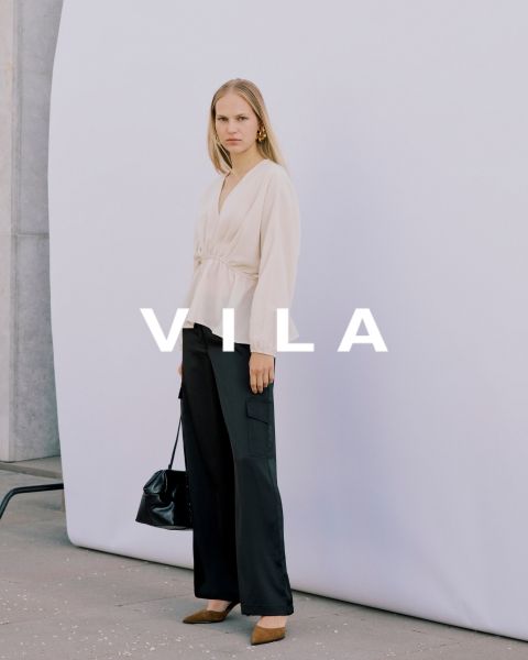 Cinco mujeres en ropa casual, incluyendo vaqueros y tops en colores neutros, están de pie juntas y sonriendo frente a un fondo sencillo. El texto superpuesto dice "Vila Clothes".