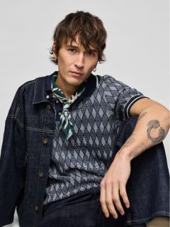 Un joven con chaqueta y pantalones vaqueros está arrodillado y mirando hacia un lado. Lleva zapatillas blancas y tiene un tatuaje visible en el antebrazo. El fondo es de color gris claro uniforme.