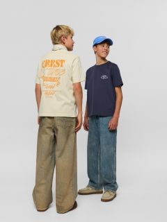 Dos chicos en vaqueros y camisas vaqueras posan juntos. Uno está sentado en un taburete con las manos en el regazo y una bufanda alrededor del cuello, mientras que el otro está de pie con el brazo en el taburete y lleva un gorro.