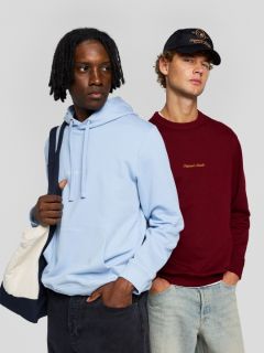 Un joven con cabello castaño corto, que lleva una camiseta color crema con un estampado azul marino en la espalda, jeans azul claro y zapatillas de deporte color beige, está sentado en el suelo mirando por encima del hombro a la cámara.