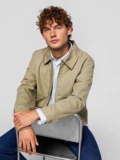 Un joven que lleva una camiseta negra con la inscripción "Original Studio", un collar plateado, una pulsera plateada y una gorra de béisbol a rayas color crema y negro, está de pie frente a un fondo liso de color gris claro.