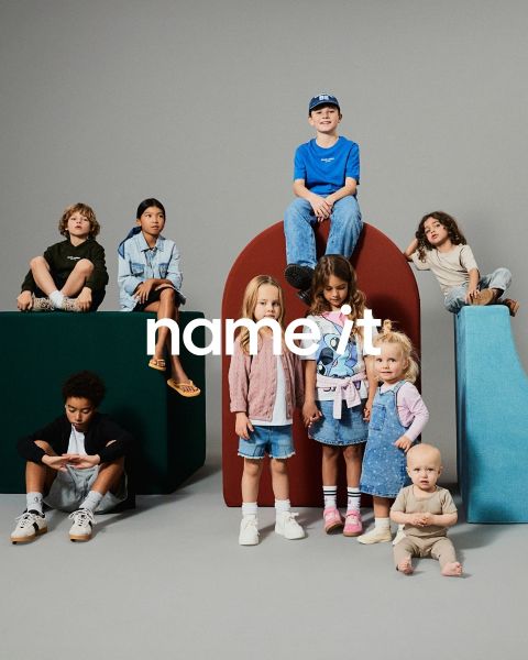 Un grupo de ocho niños diferentes están juntos frente a un fondo gris, todos vestidos con atuendos casuales a juego en colores neutros y pastel. El texto "name it" aparece en el centro de la imagen.
