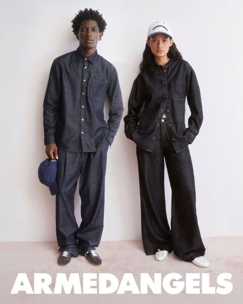 Dos jóvenes adultos vestidos con ropa vaquera están juntos al aire libre, uno con el brazo levantado, el otro con una maleta cubierta de pegatinas. En la parte inferior, en letras blancas y negrita, dice "ARMEDANGELS".