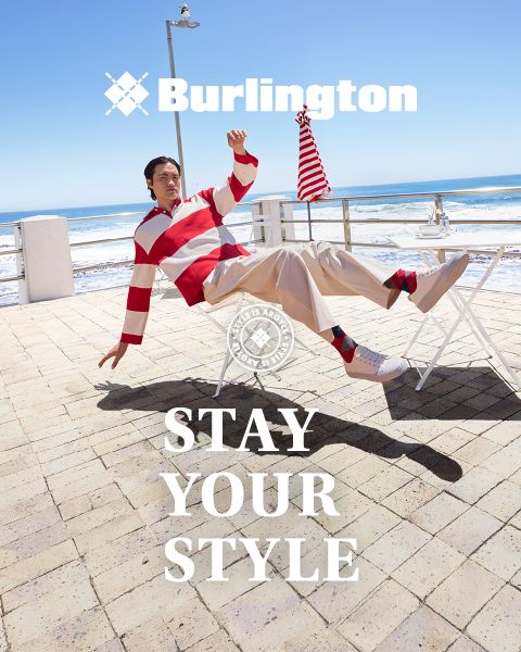 Un hombre y una mujer con atuendos elegantes posan con confianza en un banco blanco frente a una pared verde con los textos "Burlington" y "Stay Your Style" encima y debajo de ellos.