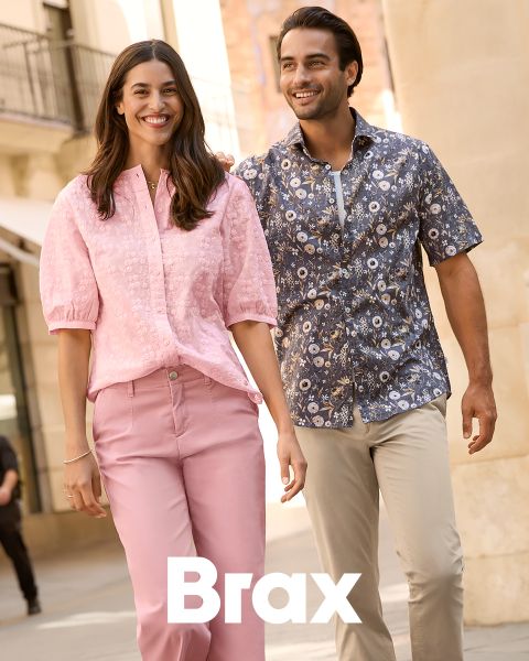 Una mujer con un abrigo estampado y un suéter rojo está de pie junto a un hombre con un abrigo de invierno azul marino y pantalones beige. Están apoyados en una pared de piedra al aire libre. La palabra "Brax" aparece en la parte inferior de la imagen.