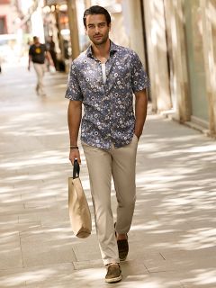 Un hombre con cabello corto y oscuro y barba está apoyado en una barandilla de piedra al aire libre. Lleva una chaqueta a cuadros roja y blanca sobre un suéter color crema y jeans azules. En el fondo se ven edificios urbanos.