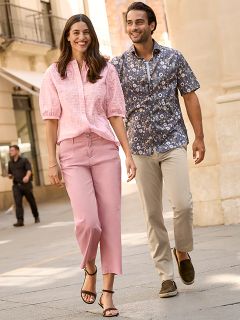 Una mujer con un abrigo de piel estampado y una blusa roja está de pie sonriendo junto a un hombre con una chaqueta de invierno azul marino y un suéter beige. Están de pie afuera junto a una pared de piedra, y la mujer sostiene un bolso marrón.