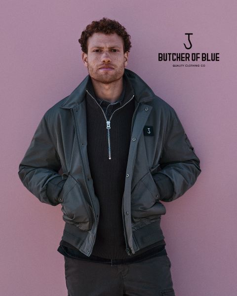 Un hombre con cabello rizado lleva una chaqueta verde oscuro sobre un suéter con cremallera y está de pie frente a un fondo rosa. El logo "Butcher of Blue Quality Clothing Co" se ve en la esquina superior derecha.