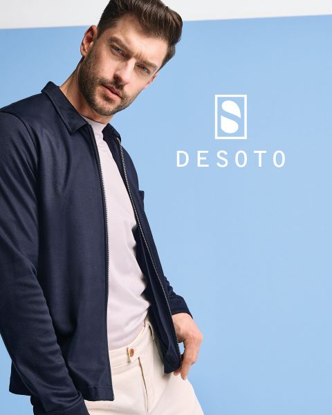Un hombre con una camisa estampada de manga larga y pantalones oscuros está de pie frente a un fondo gris. Mira hacia un lado con las manos en los bolsillos. La inscripción "DESOTO" y el logotipo de DESOTO se ven en la parte inferior.