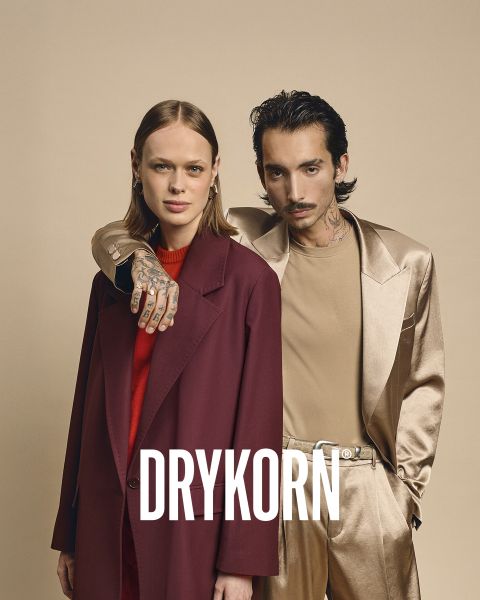 Una elegante mujer mayor con cabello rubio platino y un abrigo de piel negro está sentada en un sofá antiguo junto a un perro grande marrón. La palabra "DRYKORN" se puede leer en la parte inferior de la imagen. La iluminación cálida crea una atmósfera lujosa.