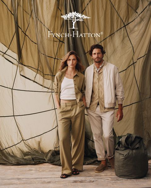 Un hombre con cabello castaño ondulado y barba lleva un jersey azul marino y está de pie frente a una pared beige con textura. El logo y el nombre de Fynch-Hatton aparecen en la esquina superior derecha.