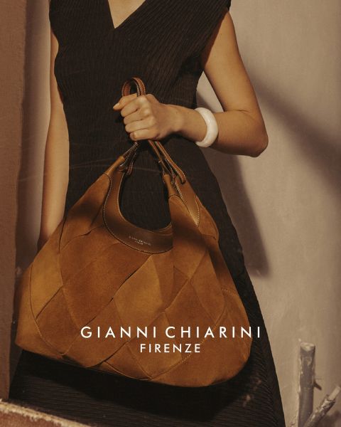 Persona con una gabardina larga de cuero negra y gafas de sol, sosteniendo un bolso pequeño con estampado de leopardo. El fondo es gris, con el texto "GIANNI CHIARINI FIRENZE" en blanco sobre la imagen.