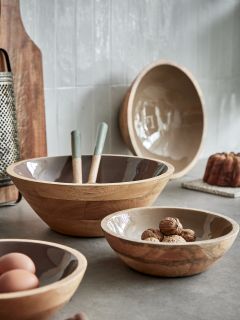 Sobre una mesa de cocina hay tres cuencos de madera de diferentes tamaños. En un cuenco hay nueces, en otro dos huevos y en el más grande hay utensilios. En el fondo se ven un rallador, una tabla de cortar y un cuenco fijado a la pared.